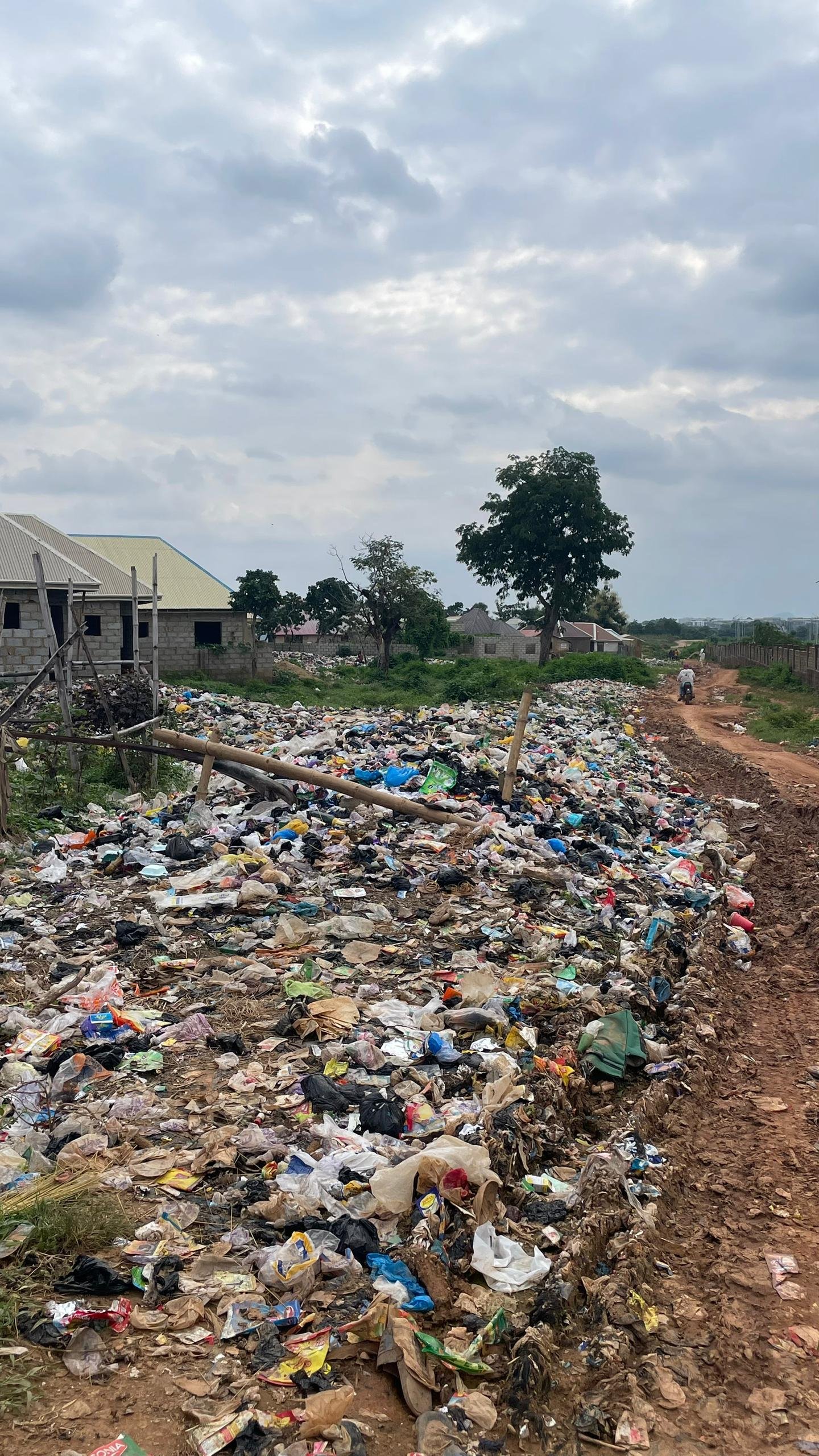Open waste dump at the entrance of Karonmajiji community in Abuja, showing heaps of refuse scattered near homes and pathways.