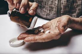 A close-up illustration of an elderly Nigerian woman holding prescription pills, with a faded pharmacy counter in the background — symbolising the high cost of survival