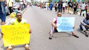 Nigerian university students sit on the floor holding placards that read “End ASUU Strikes Now” during protest over prolonged university closures.